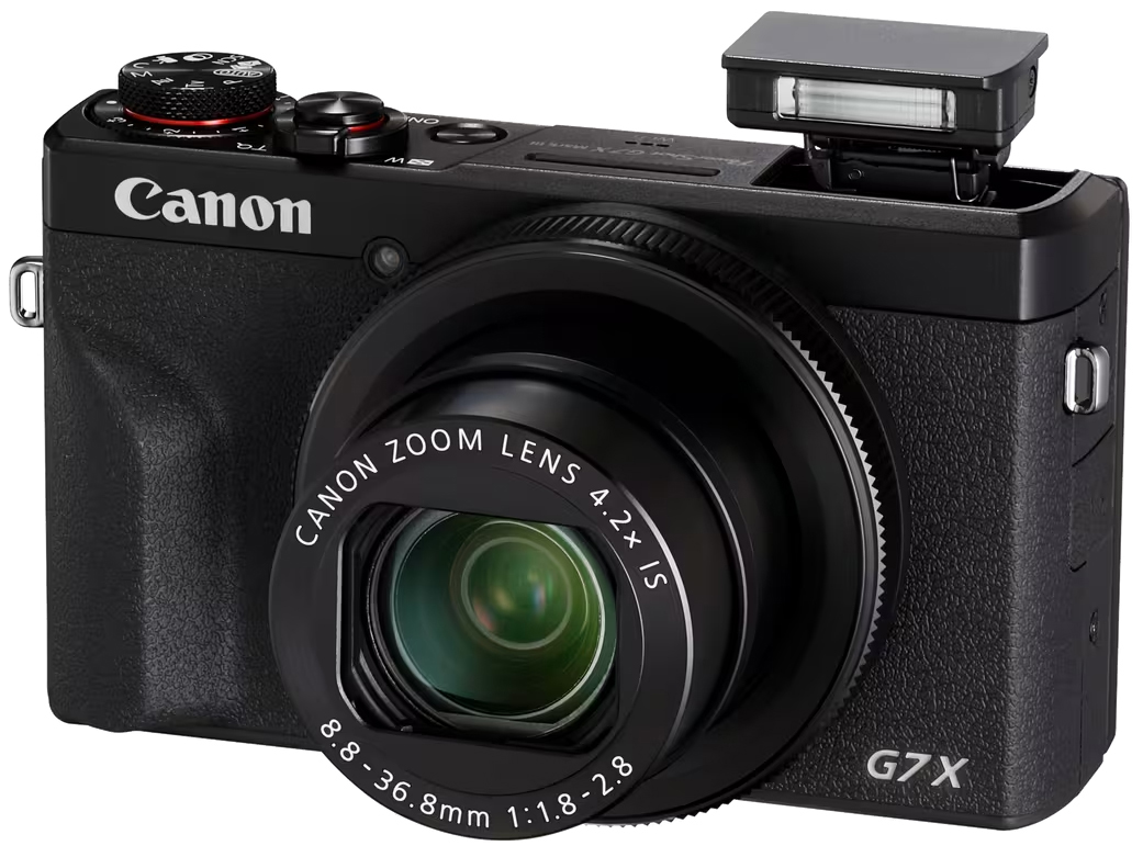 Czarny aparat Canon PowerShot G7X Mark III widoczny pod kątem na białym tle. Model ma wysunięty tubus obiektywu z białymi oznaczeniami oraz podniesioną, niewielką lampę błyskową typu pop-up po prawej stronie. Na fakturowanej obudowie widoczne są logotypy CANON i G7X oraz górne pokrętła z czerwonym akcentem.