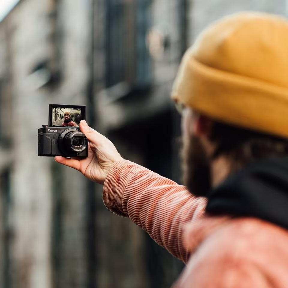 Czarny aparat Canon PowerShot G7X Mark III trzymany w wyciągniętej ręce wykonuje selfie. Ekran aparatu jest maksymalnie odchylony pionowo do góry o 180 stopni, pokazując twarz mężczyzny w żółtej czapce. Tło miejskiej ulicy jest zamazane.