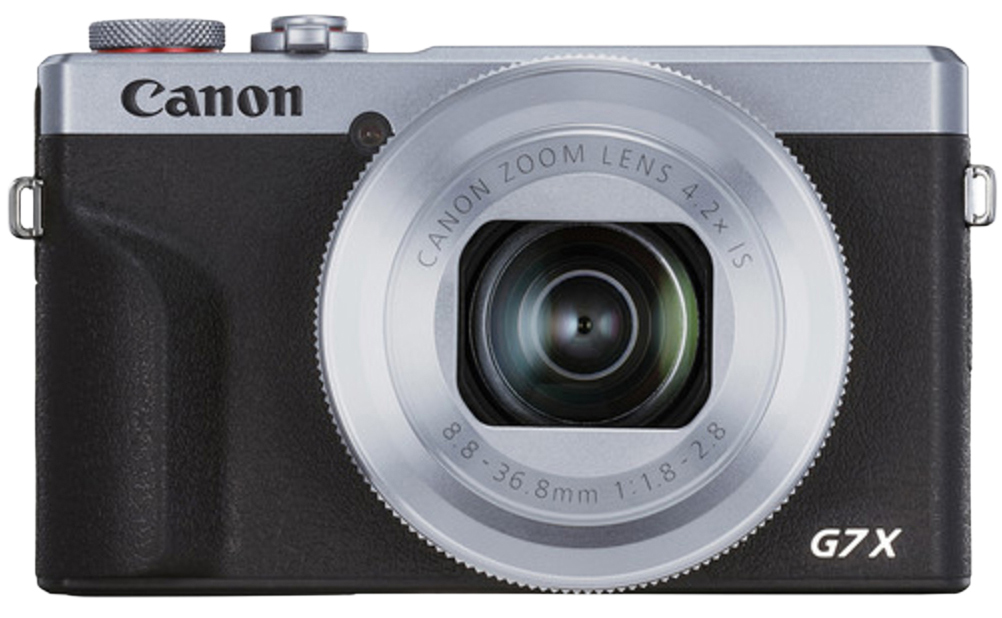 Srebrny aparat kompaktowy Canon PowerShot G7X Mark III na białym tle, widok od przodu. Korpus ma chropowatą strukturę z białym logo CANON oraz wyraźnym oznaczeniem modelu G7X. Centralne miejsce zajmuje duży obiektyw zmiennoogniskowy z widocznymi parametrami optycznymi oraz czerwonym akcentem przy górnym pokrętle.