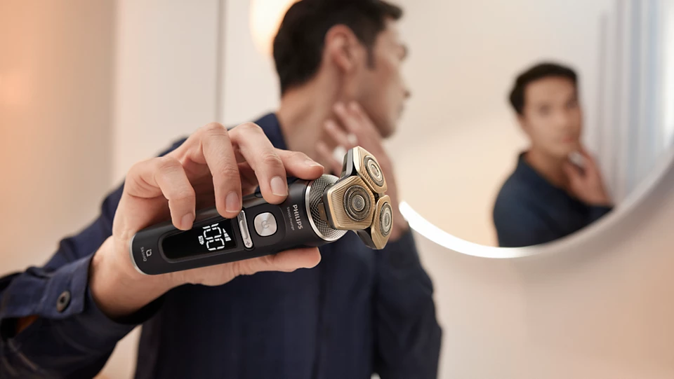 Mężczyzna w ciemnej koszuli trzyma golarkę elektryczną Philips S9000 Prestige SP9840/32 z wyświetlaczem "100%" na pierwszym planie, prezentując produkt. W tle, odbicie mężczyzny w lustrze, gdzie dotyka on swojej szyi. Scena rozgrywa się w jasnej łazience z delikatnym oświetleniem, tworząc intymną atmosferę.