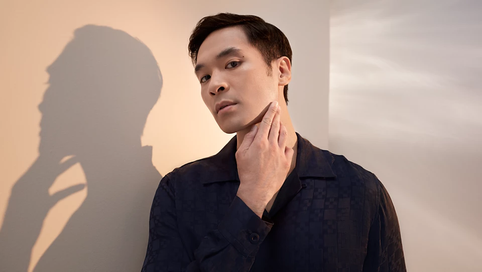 Mężczyzna w ciemnej koszuli z krótkim rękawem, patrzący wprost na widza. Jego prawa dłoń dotyka delikatnie szyi. Tło stanowi jasna ściana, na którą pada cień z lewej strony, tworząc rozmyty kształt dłoni.