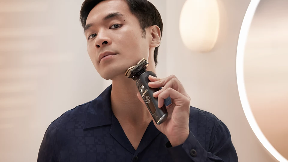 Mężczyzna w ciemnej koszuli goli się golarką elektryczną Philips S9000 Prestige SP9840/32. Urządzenie, z widocznymi trzema ruchomymi głowicami tnącymi w kolorze srebrno-złotym, jest przyłożone do jego prawego policzka. W tle znajduje się jasna łazienka z rozmyciem tła.