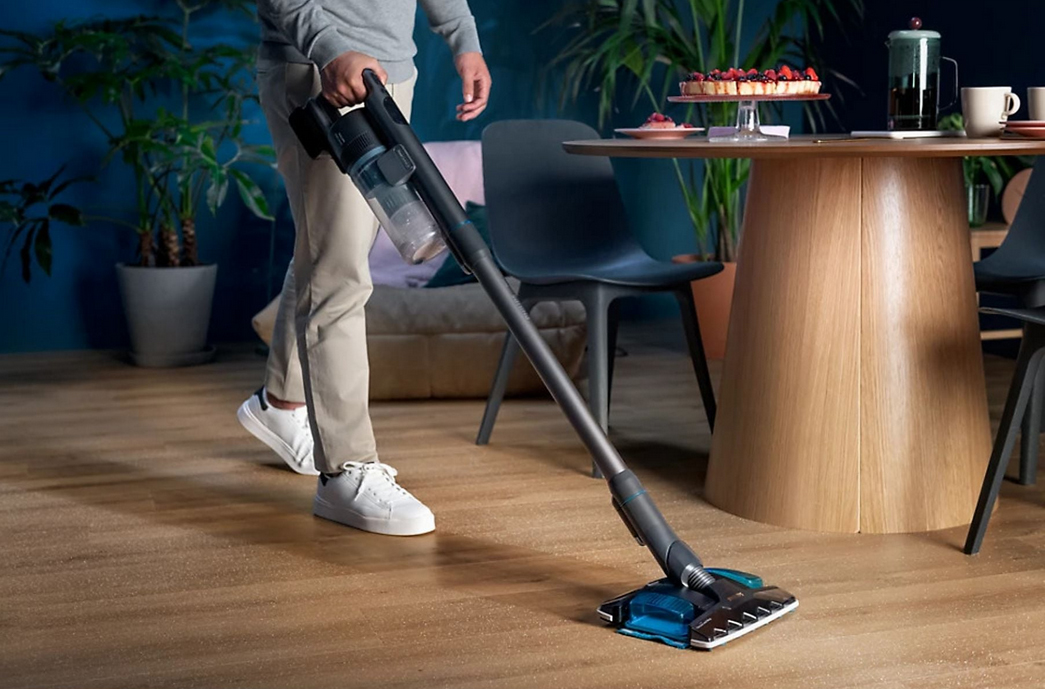Osoba odkurza i mopuje drewnianą podłogę odkurzaczem Philips SpeedPro 8000 XC8157/01 w nowoczesnym salonie. Urządzenie w kolorze ciemnoszarym posiada ssawkę z turkusowymi elementami i niebieską nakładką. W tle widoczny jest stół z poczęstunkiem oraz dekoracyjne rośliny, co podkreśla domowy kontekst wykorzystania sprzętu.